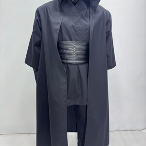 Op de afbeelding: Een zwart kostuum met capuchon en bijpassende zwarte riem. Het kostuum bevat een lange, vloeiende jurk, een tuniek en een broek. De riem heeft zilverkleurige klinknagels. Het kostuum wordt op een mannequin getoond.