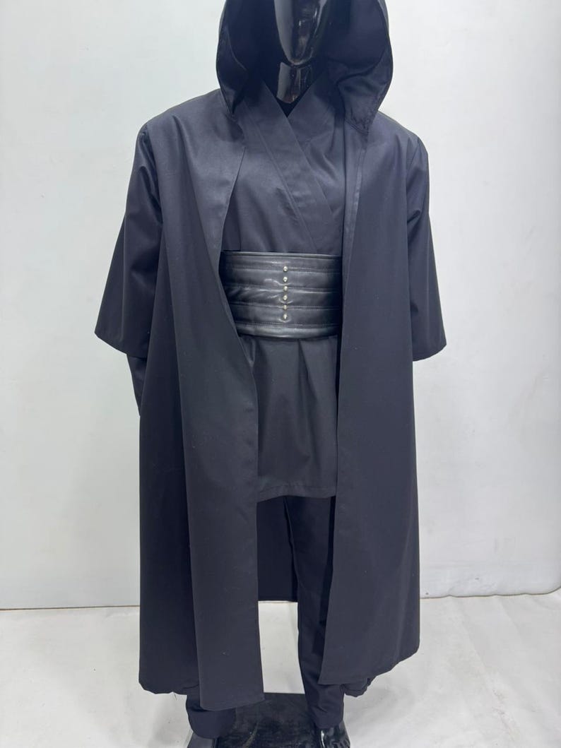 Op de afbeelding: Een zwart kostuum met capuchon en bijpassende zwarte riem. Het kostuum bevat een lange, vloeiende jurk, een tuniek en een broek. De riem heeft zilverkleurige klinknagels. Het kostuum wordt op een mannequin getoond.