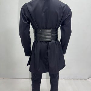 Op de afbeelding: Zwart vechtsportuniform met een brede, zwarte riem. Het uniform bestaat uit een jas met lange mouwen en een bijpassende broek. De riem is gemaakt van een dik, getextureerd materiaal. De outfit wordt gepresenteerd op een mannequin.
