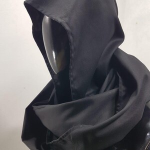 May include: A black hooded cloak made of a smooth, matte fabric. The hood is deep and the cloak drapes around the shoulders. The cloak is displayed on a black mannequin head.