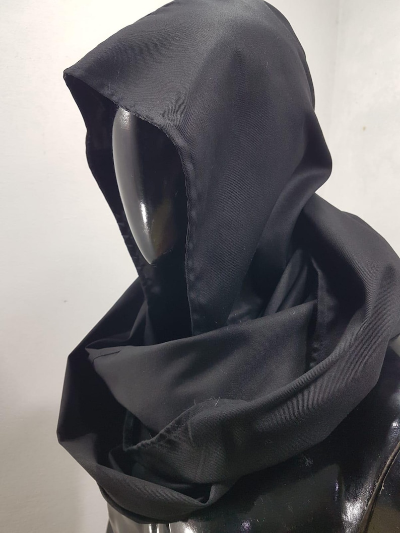 May include: A black hooded cloak made of a smooth, matte fabric. The hood is deep and the cloak drapes around the shoulders. The cloak is displayed on a black mannequin head.
