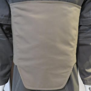Puede incluir: Un chaleco protector de color beige usado sobre un uniforme gris oscuro. El chaleco es rectangular con un borde inferior curvo y est&aacute; hecho de un material liso, posiblemente acolchado. El uniforme tiene mangas largas y costuras visibles.