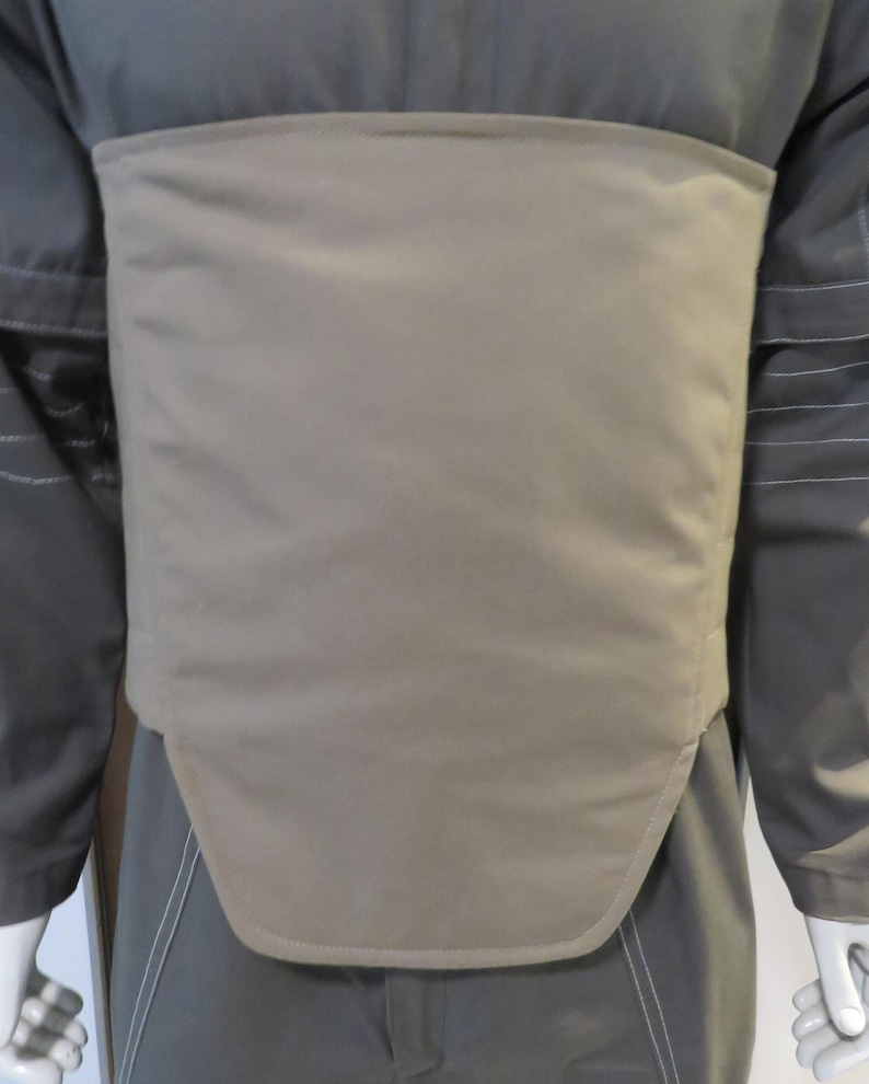 Puede incluir: Un chaleco protector de color beige usado sobre un uniforme gris oscuro. El chaleco es rectangular con un borde inferior curvo y est&aacute; hecho de un material liso, posiblemente acolchado. El uniforme tiene mangas largas y costuras visibles.