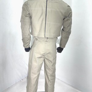 Puede incluir: Un conjunto beige de dos piezas sobre un maniqu&iacute;. La chaqueta tiene un cuello alto, una cremallera en la espalda y costuras blancas. Los pantalones son de cintura alta con costuras blancas. El conjunto se exhibe sobre un soporte negro.