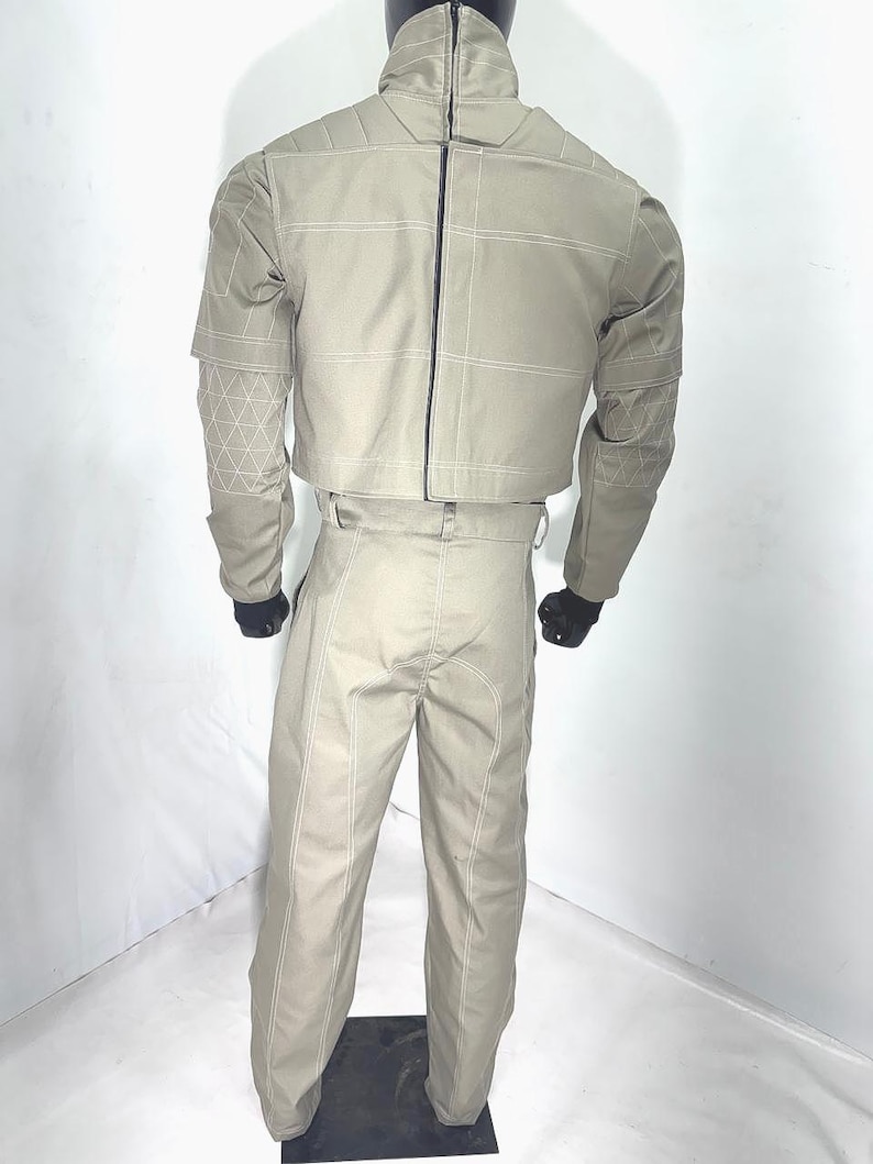 Puede incluir: Un conjunto beige de dos piezas sobre un maniqu&iacute;. La chaqueta tiene un cuello alto, una cremallera en la espalda y costuras blancas. Los pantalones son de cintura alta con costuras blancas. El conjunto se exhibe sobre un soporte negro.