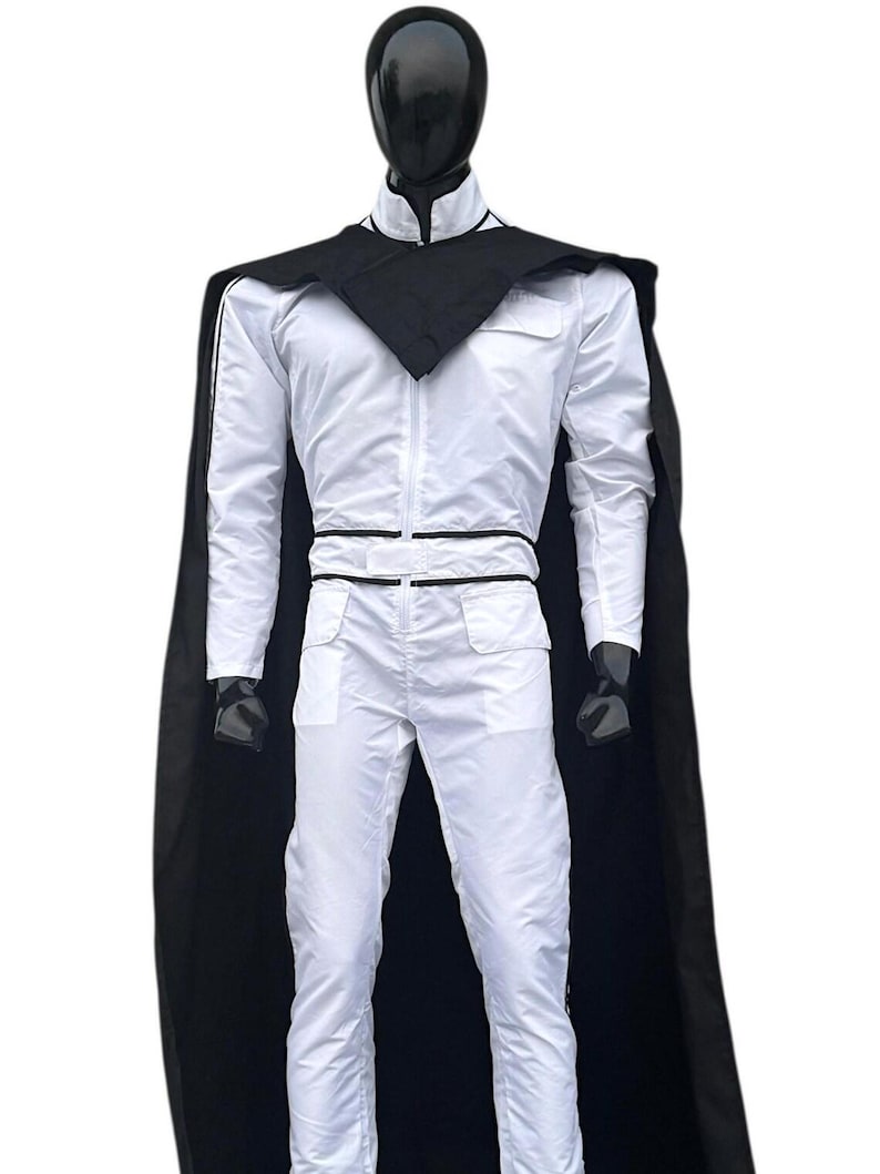 May include: A white jumpsuit with a black cape and collar is displayed on a mannequin. The jumpsuit has a zip-up front, pockets, and black trim. The black cape drapes over the shoulders, creating a striking contrast.