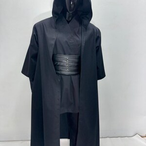 Op de afbeelding: Een zwart kostuum met capuchon en bijpassende outfit, gepresenteerd op een mannequin. Het kostuum omvat een lange, vloeiende mantel, een tuniek, een broek en een brede riem met metalen accenten. Ideaal voor cosplay of kostuumevenementen.