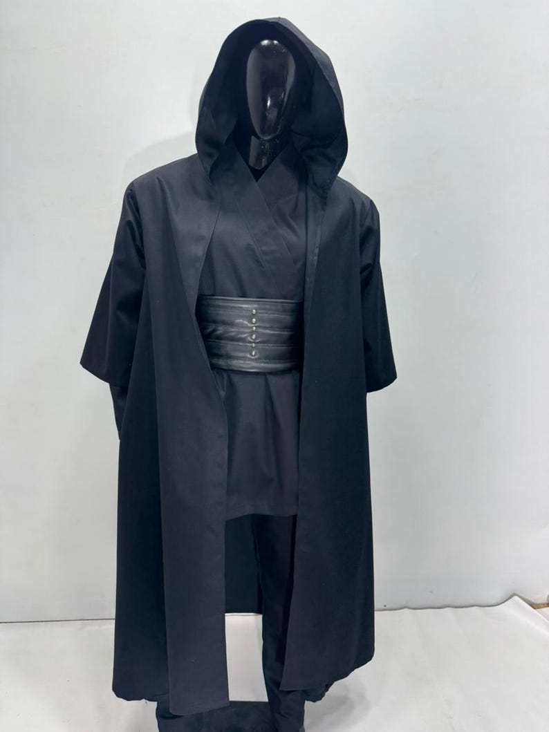 Op de afbeelding: Een zwart kostuum met capuchon en bijpassende outfit, gepresenteerd op een mannequin. Het kostuum omvat een lange, vloeiende mantel, een tuniek, een broek en een brede riem met metalen accenten. Ideaal voor cosplay of kostuumevenementen.
