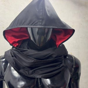 May include: Black hooded cowl with a red satin lining. The cowl is draped over a mannequin, showcasing the design. The hood is pointed and the cowl wraps around the neck and shoulders.