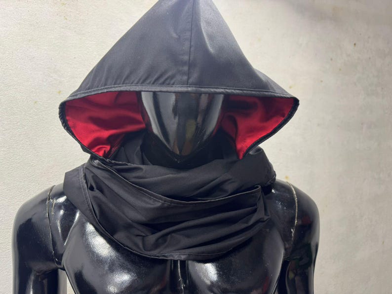 May include: Black hooded cowl with a red satin lining. The cowl is draped over a mannequin, showcasing the design. The hood is pointed and the cowl wraps around the neck and shoulders.