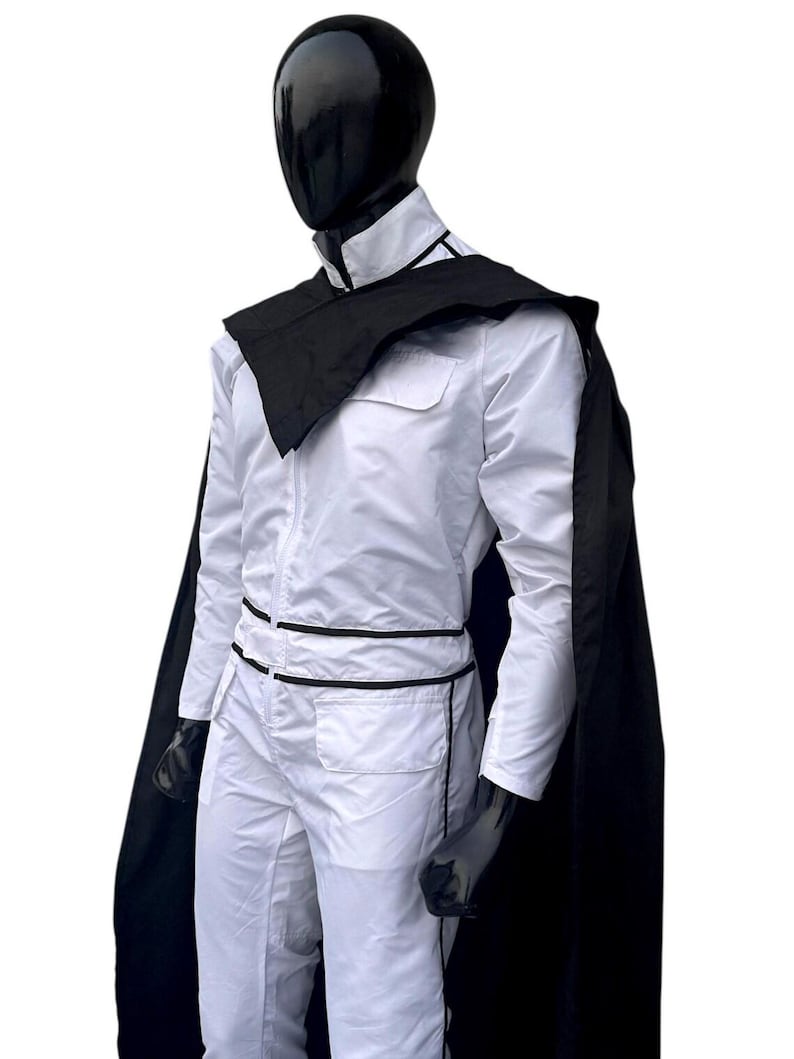 May include: A white jumpsuit with black trim and a matching black cape. The outfit is displayed on a mannequin with a black head and gloves. The jumpsuit has a zip-up front and pockets.