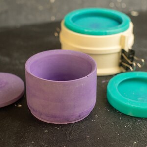 May include: Three round molds for making concrete planters. The molds are purple, white, and teal. The teal mold is on a black surface.