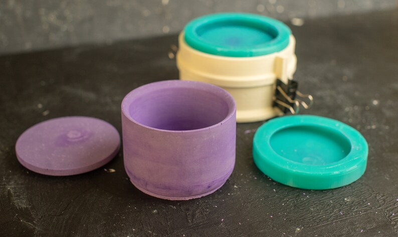 May include: Three round molds for making concrete planters. The molds are purple, white, and teal. The teal mold is on a black surface.
