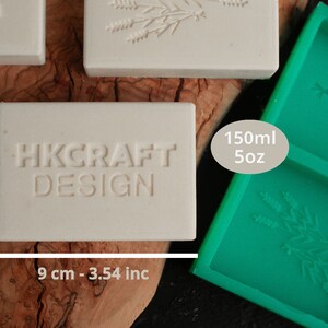 May include: A white rectangular soap bar with the text "HKCRAFT DESIGN" embossed on it. The soap bar is 6.5 inches tall and 3.54 inches wide. A green silicone mold with two rectangular cavities is shown in the background. The mold is labeled "150ml 5oz".