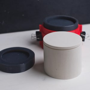 May include: Three concrete containers with lids. One container is white with a white lid, one is red with a black lid, and one is black with a black lid.
