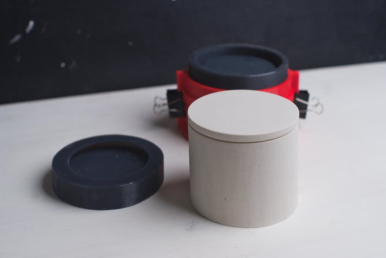May include: Three concrete containers with lids. One container is white with a white lid, one is red with a black lid, and one is black with a black lid.