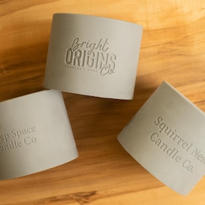 May include: Three gray candle jars with different company names embossed on the front. The jars are on a light brown wooden surface. The names are "Bright Origins Candle Co.", "Deep Space Candle Co.", and "Squirrel Nest Candle Co."