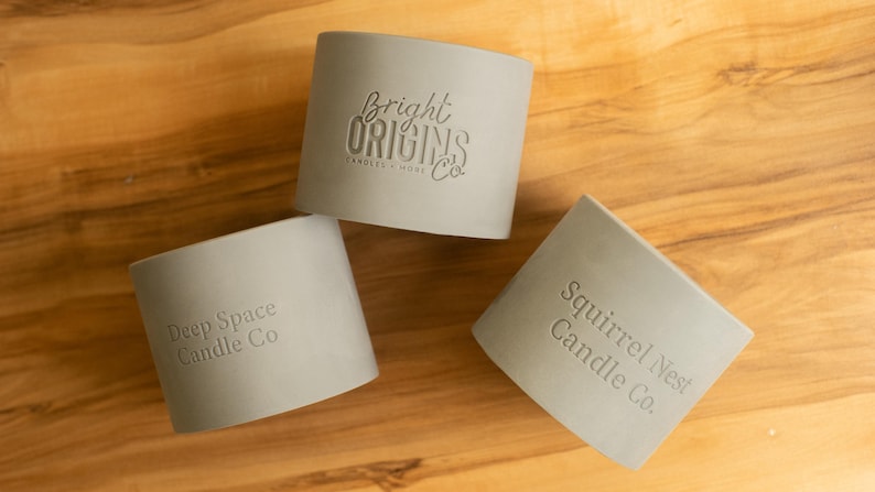May include: Three gray candle jars with different company names embossed on the front. The jars are on a light brown wooden surface. The names are "Bright Origins Candle Co.", "Deep Space Candle Co.", and "Squirrel Nest Candle Co."