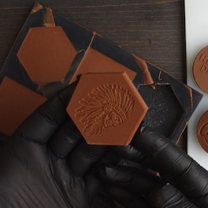 Può includere: Una caramella al cioccolato esagonale marrone con un disegno a rilievo di un copricapo nativo americano. La caramella è tenuta da una mano guantata, con uno stampo scuro visibile sullo sfondo. Sono visibili anche altre caramelle.