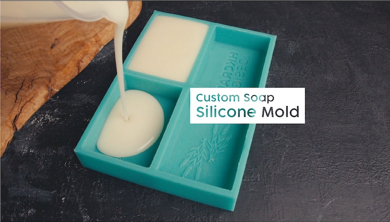May include: A teal blue silicone soap mold with three compartments. The mold has a rectangular shape with a rounded edge and a design of wheat stalks on the bottom. The mold is being filled with a white liquid.