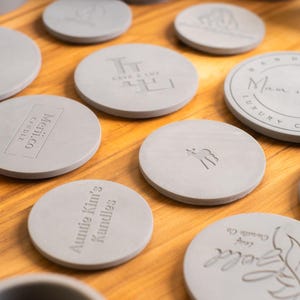 May include: A collection of round, gray, concrete candle lids with various text and designs. The lids are arranged on a wooden surface. Some of the designs include "Auntie Kim's Candles", "Lumen Soy Candles", and "Man & Pax Luxury Comfort".
