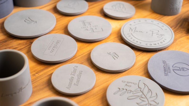 May include: A collection of round, gray, concrete candle lids with various text and designs. The lids are arranged on a wooden surface. Some of the designs include "Auntie Kim's Candles", "Lumen Soy Candles", and "Man & Pax Luxury Comfort".