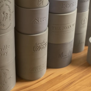 May include: A collection of gray and blue silicone candle holders with various embossed designs, including "Candle Cozy", "Bright Origins", "The Joan W", and "bonita".