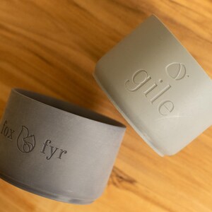 May include: Two gray silicone cup lids with different logos. The lid on the left has a fox and the text "fox fyr". The lid on the right has the text "gile" and a small acorn.