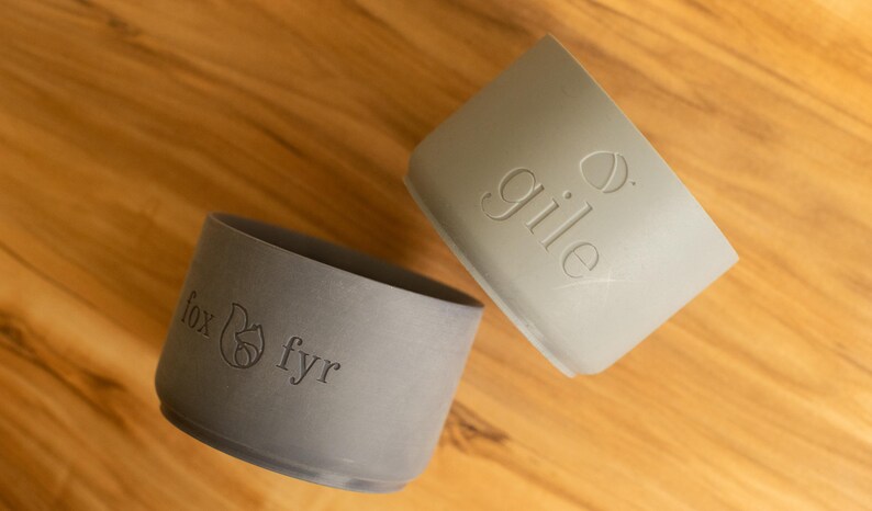 May include: Two gray silicone cup lids with different logos. The lid on the left has a fox and the text "fox fyr". The lid on the right has the text "gile" and a small acorn.