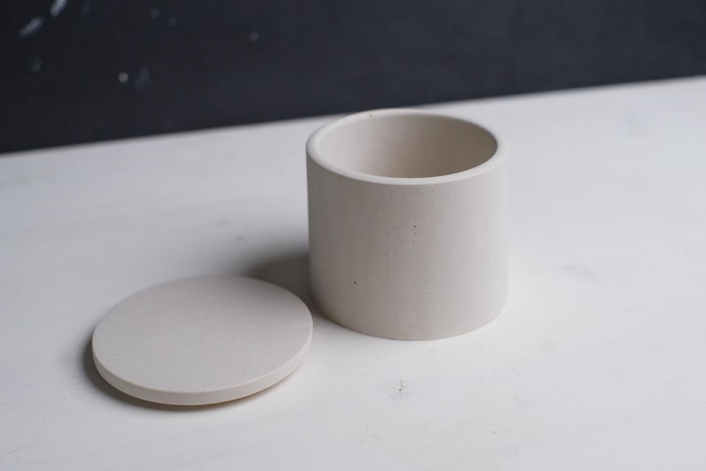 May include: A white cylindrical container with a matching lid. The container is made of a textured material, possibly concrete or ceramic.