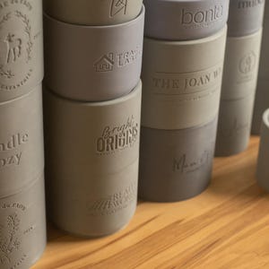 May include: A collection of gray and blue silicone candle holders with various embossed designs, including "Candle Cozy", "Bright Origins", "The Joan W", and "bonita".