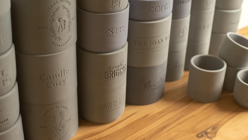 May include: A collection of gray and blue silicone candle holders with various embossed designs, including "Candle Cozy", "Bright Origins", "The Joan W", and "bonita".