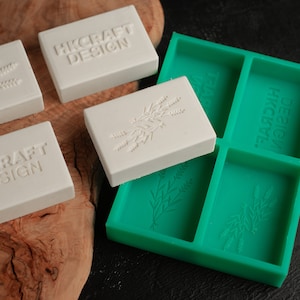 May include: Four white soap bars with the text "HKcraft Design" and a floral design. The soap bars are sitting on a wooden surface next to a green silicone mold with the same floral design.