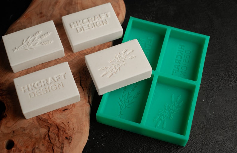 May include: Four white soap bars with the text "HKcraft Design" and a floral design. The soap bars are sitting on a wooden surface next to a green silicone mold with the same floral design.