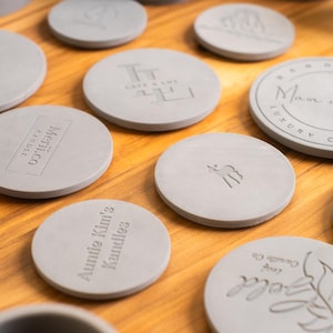May include: A collection of round, gray, concrete candle lids with various text and designs. The lids are arranged on a wooden surface. Some of the designs include "Auntie Kim's Candles", "Lumen Soy Candles", and "Man & Pax Luxury Comfort".