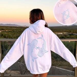May include: White hoodie with a blue outline of two whale sharks facing each other with a small heart between them. The hoodie has a drawstring hood.