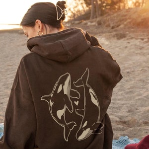May include: A brown hoodie with a graphic of two orcas swimming together. The orcas are outlined in white and have a heart shape between them.