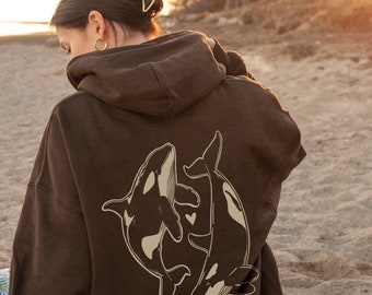 May include: A brown hoodie with a graphic of two orcas swimming together. The orcas are outlined in white and have a heart shape between them.