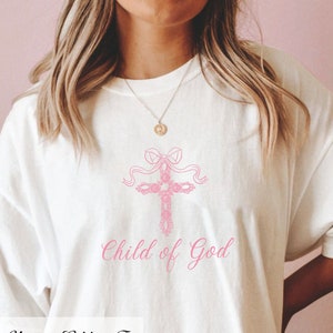 May include: White t-shirt featuring a pink floral cross design and the text "Child of God".