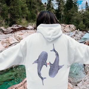 Könnte beinhalten: Weißer Hoodie mit einem Aufdruck von zwei Walhaien, die sich gegenüberstehen, mit einem kleinen roten Herzen dazwischen.
