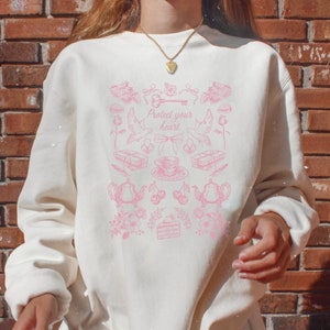 May include: A white sweatshirt with a pink graphic design featuring a key, a heart, and various other small images. The text "Protect your heart" is printed on the sweatshirt.