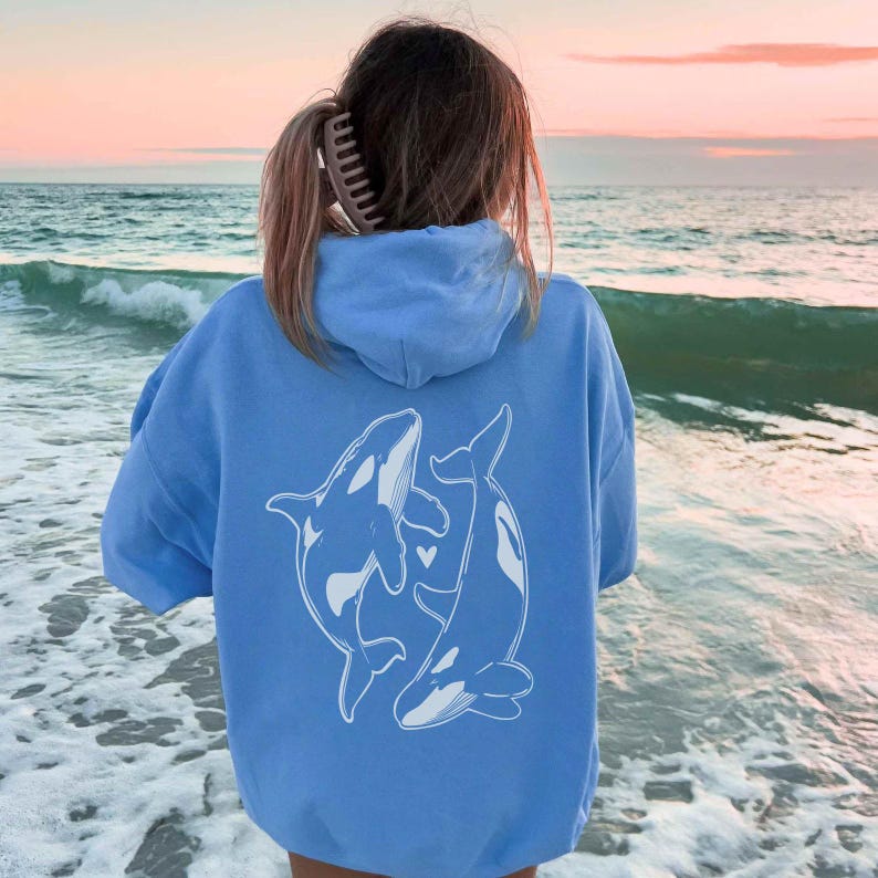 May include: A light blue hoodie with a white graphic of two orcas swimming together. The hoodie has a hood and a kangaroo pocket.