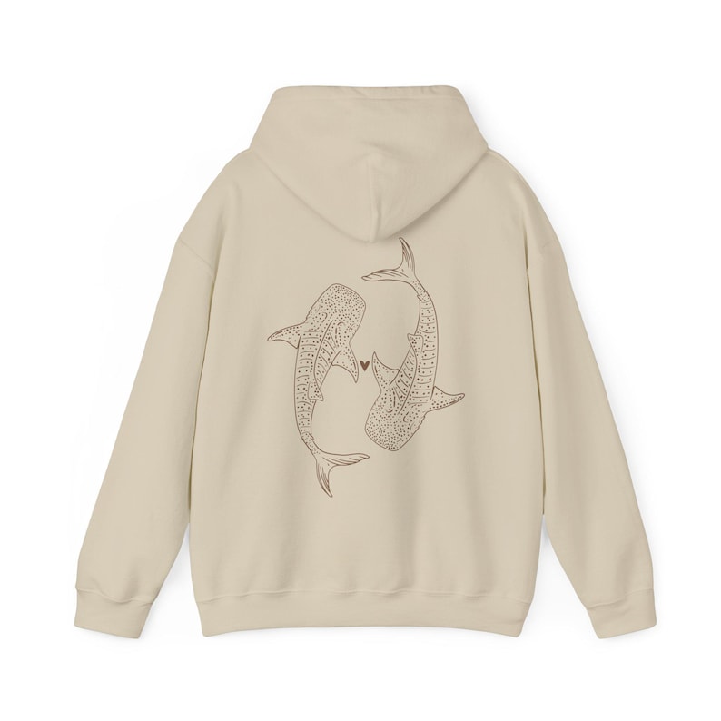 Puede incluir: Una sudadera con capucha de color crema con un dibujo lineal marr&oacute;n de dos tiburones ballena que se miran en la espalda. Los tiburones est&aacute;n dibujados con un coraz&oacute;n entre ellos.