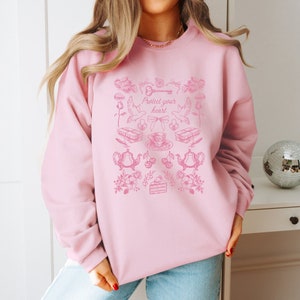 May include: Pink sweatshirt with a graphic design of a key, doves, teacups, cherries, and a cake. The text "Protect your heart" is printed on the sweatshirt.