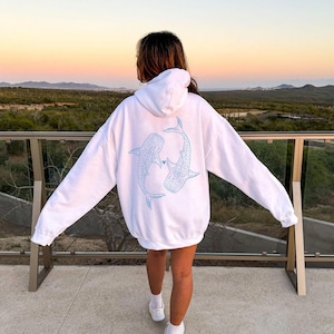 Puede incluir: Sudadera blanca con un contorno azul de dos tiburones ballena que se miran y se tocan la nariz. Los tiburones est&aacute;n delineados en azul claro y tienen un peque&ntilde;o coraz&oacute;n entre ellos.