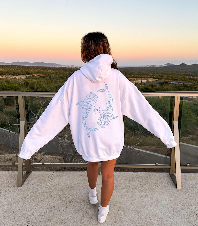 Puede incluir: Sudadera blanca con un contorno azul de dos tiburones ballena que se miran y se tocan la nariz. Los tiburones est&aacute;n delineados en azul claro y tienen un peque&ntilde;o coraz&oacute;n entre ellos.