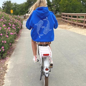 Puede incluir: Una persona montando una bicicleta blanca con una cesta rosa en la parte trasera. La persona lleva una sudadera con capucha azul real con un dise&ntilde;o de tibur&oacute;n ballena blanco en la espalda.