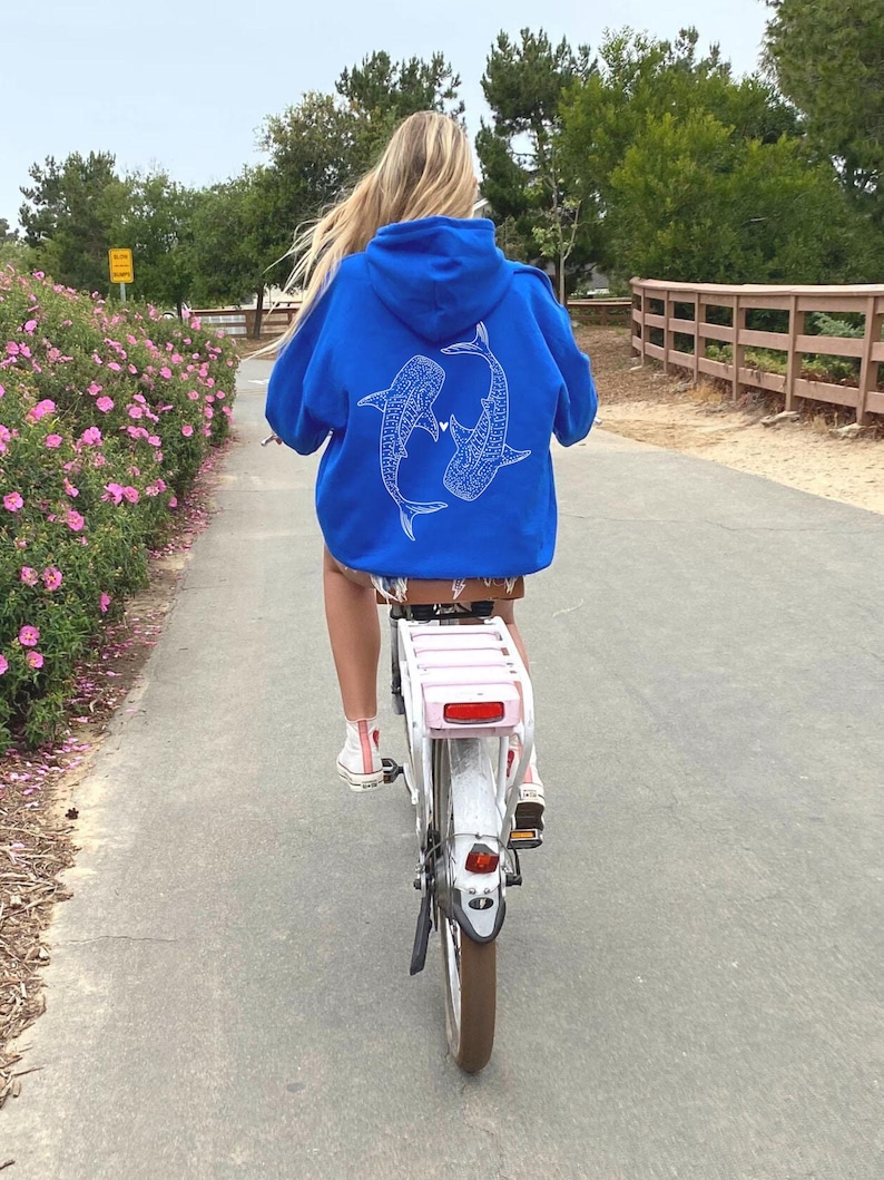 Puede incluir: Una persona montando una bicicleta blanca con una cesta rosa en la parte trasera. La persona lleva una sudadera con capucha azul real con un dise&ntilde;o de tibur&oacute;n ballena blanco en la espalda.