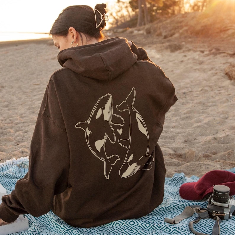 May include: A brown hoodie with a graphic of two orcas swimming together. The orcas are outlined in white and have a heart shape between them.