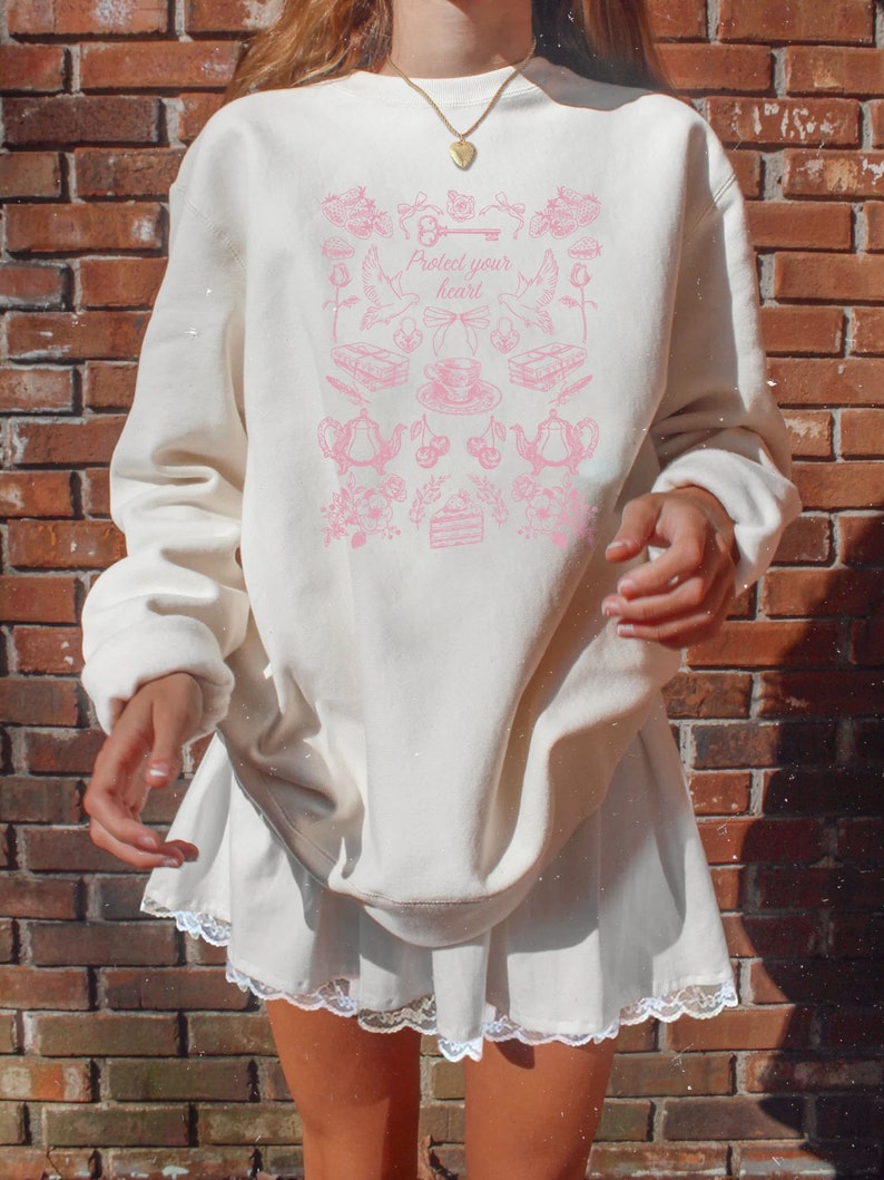 May include: A white sweatshirt with a pink graphic design featuring a key, a heart, and various other small images. The text "Protect your heart" is printed on the sweatshirt.